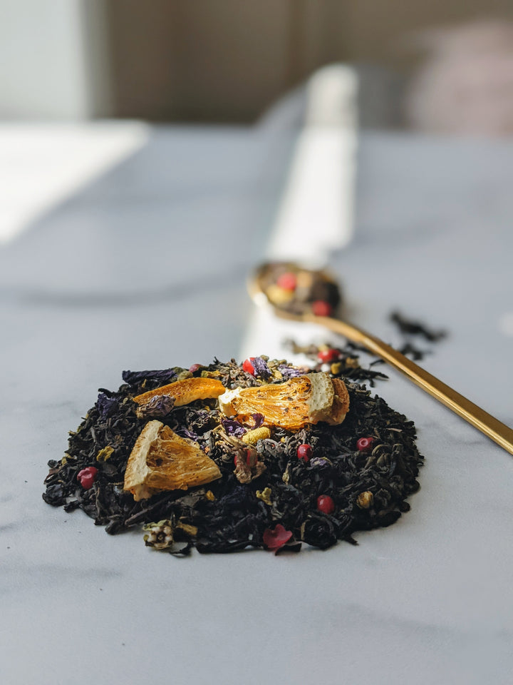 This is an image of The Silk Tea Co's 'Up in Smoke' gunpowder and Lapsang Souchong tea blend featuring pink peppercorns and orange slices. Here a small scoop of the loose leaf blend is lit by a ray of sunlight, highlighting the vibrant colors and a golden spoon featuring some of the blend.