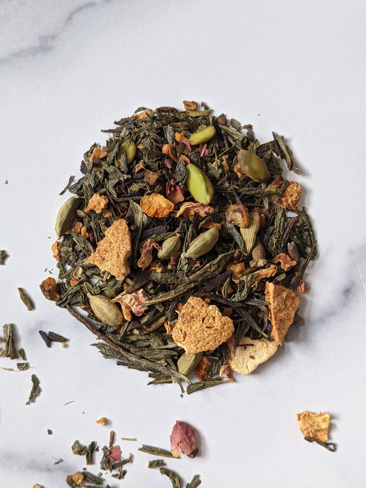 This is an image of a scoop of The Silk Tea Co's 'Eyes of Green' tea blend with cardamom, pistachios, lemon peels, and pink yellow peels in view with a mix of green and black tea leaves., 