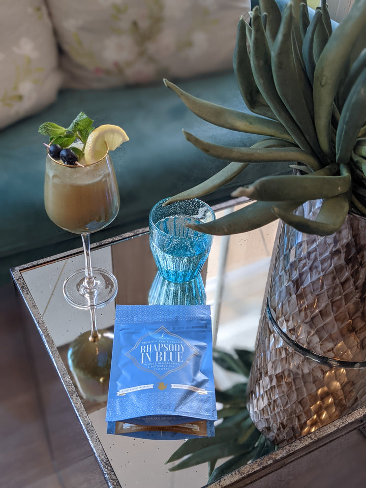 The Silk Tea Co features a blue colored loose leaf tea pouch with the name 'Rhapsody in Blue' on a mirrored table with a cocktail glass and pretty blue water glass next to a succulent planter.