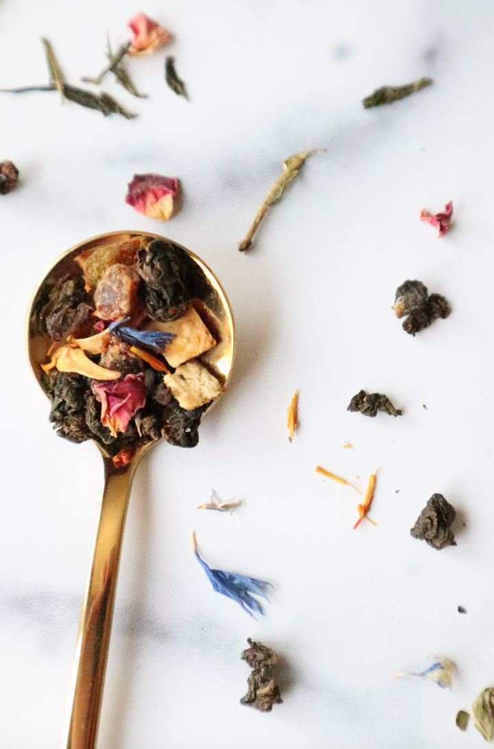 This is a gold spoonful of our Morning Sex tea blend set against a marble countertop with loose leaf tea pieces sprinkled like confetti all around.